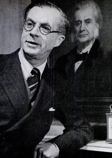 Black-and-white photograph of Julian Huxley in round glasses and a pinstriped suit, mid-speech, in front of a painted portrait of his grandfather Thomas Henry Huxley.